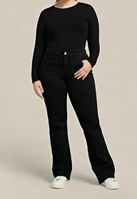 Femme portant un haut noir à manches longues, un pantalon noir taille haute et des baskets blanches, debout devant un fond uni.