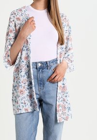 Květinové kimono s bílým základem, modrými a růžovými květinovými vzory, lehký materiál, volný střih, oversized rukávy, nošené přes bílé tílko a džíny.