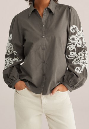 Femme portant une chemise grise boutonnée avec une broderie en dentelle blanche sur les manches et un pantalon clair, mains dans les poches.