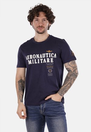 T-shirt in cotone blu navy con testo bianco "AERONAUTICA MILITARE", emblema dorato sul petto e toppe sul maniche sinistra. Collo rotondo.