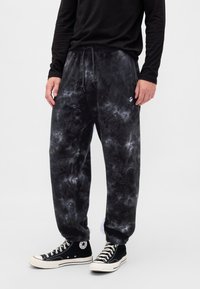 Czarne spodnie dresowe z efektem tie-dye posiadają elastyczną talię ze sznurkiem, boczne kieszenie oraz mały akcent w postaci logo. Połączone z czarnymi sneakersami.