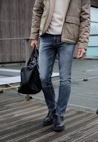 Světle modré džínové kalhoty s úzkým střihem. Kombinovány s béžovým svetrem, hnědou bundou a černými koženými kotníkovými botami. Nese černou kabelku.