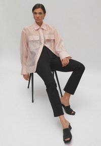 Translucentes Blush-Shirt mit zwei vorderen Taschen, kombiniert mit schwarzen Slim-Pants und strukturierten schwarzen Mules. Minimalistisches Design, lockere Passform.