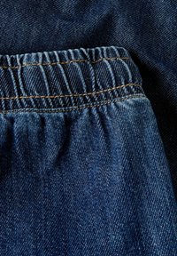 Tumši zils džinsu audums ar savāktu elastīgu vidukļa jostu. Tekstūra izskatās gluda ar redzamu šuvju detaļu vidukļa apvidū.