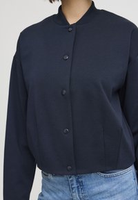 Veste cropped bleu marine en tissu lisse, avec un col rond, des boutons sur le devant et des manches longues. Associée à un jean en denim clair.