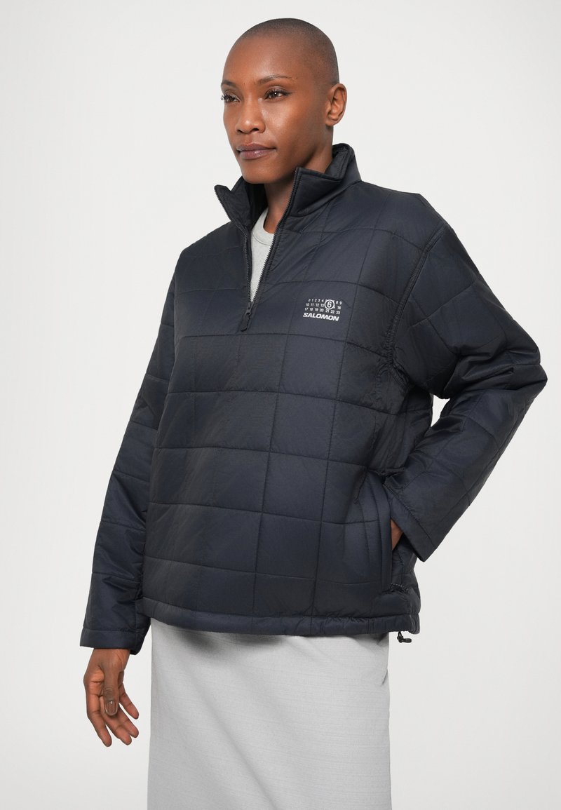 Person in einer schwarzen, gesteppte Salomon Pulloverjacke mit halbem Reißverschluss, steht mit einer Hand in der Tasche und trägt einen hellgrauen Rock vor einem weißen Hintergrund.