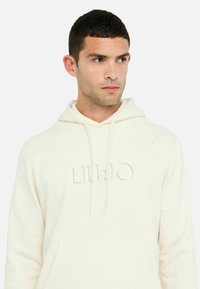 Cremefarvet hoodie lavet af blødt stof, med en lomme foran, snoretræk i hætten og præget "LIU JO" logo. Enkel, minimalistisk design.