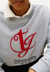 Sudadera gris con el logo rojo "TJ", texto blanco y puños acanalados. Presenta una camisa color beige debajo con cuello. Los detalles incluyen un pequeño logo en la manga.