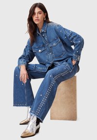Giacca di jeans e pantaloni a gamba ampia adornati con borchie argentate, di un blu medio, abbinati a stivaletti metallici argento e oro.