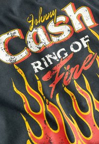 Černé tričko s odvážnými, opotřebovanými grafikami, na kterých je napsáno "Johnny Cash" bílou a červenou barvou a "RING OF FIRE" červeně, obklopené žlutými plameny.