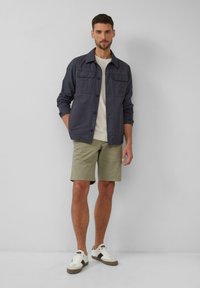 Donkerblauwe shirtjacket met borstzakken, gecombineerd met lichtgroene shorts. Model draagt witte sneakers met bruine accenten. Eenvoudig en functioneel ontwerp.