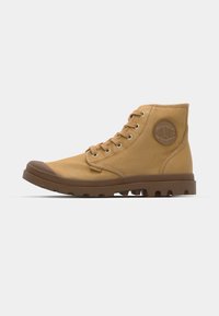 Botas de tobillo de lona color beige con una suela de goma marrón, punta redonda, ojales metálicos y una etiqueta de logo en el lateral. Presentan un tejido texturizado y cordones.