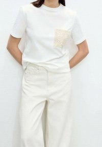 T-shirt blanc à manches courtes et col rond ; dispose d'une poche en dentelle beige. Associé à un pantalon blanc taille haute.