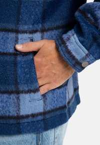 Fleece jacket in shades of blue with a plaid pattern, featuring a side pocket and snap button detail on the cuff. Smooth, textured fabric.
