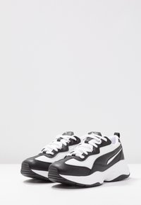 Musta-valkoiset urheilukengät, joissa on teksturoitu nahkainen päällinen, verkkokangasosat ja valkoiset nauhat. Varustettu paksulla valkoisella pohjalla ja takana olevalla vetolenkillä.