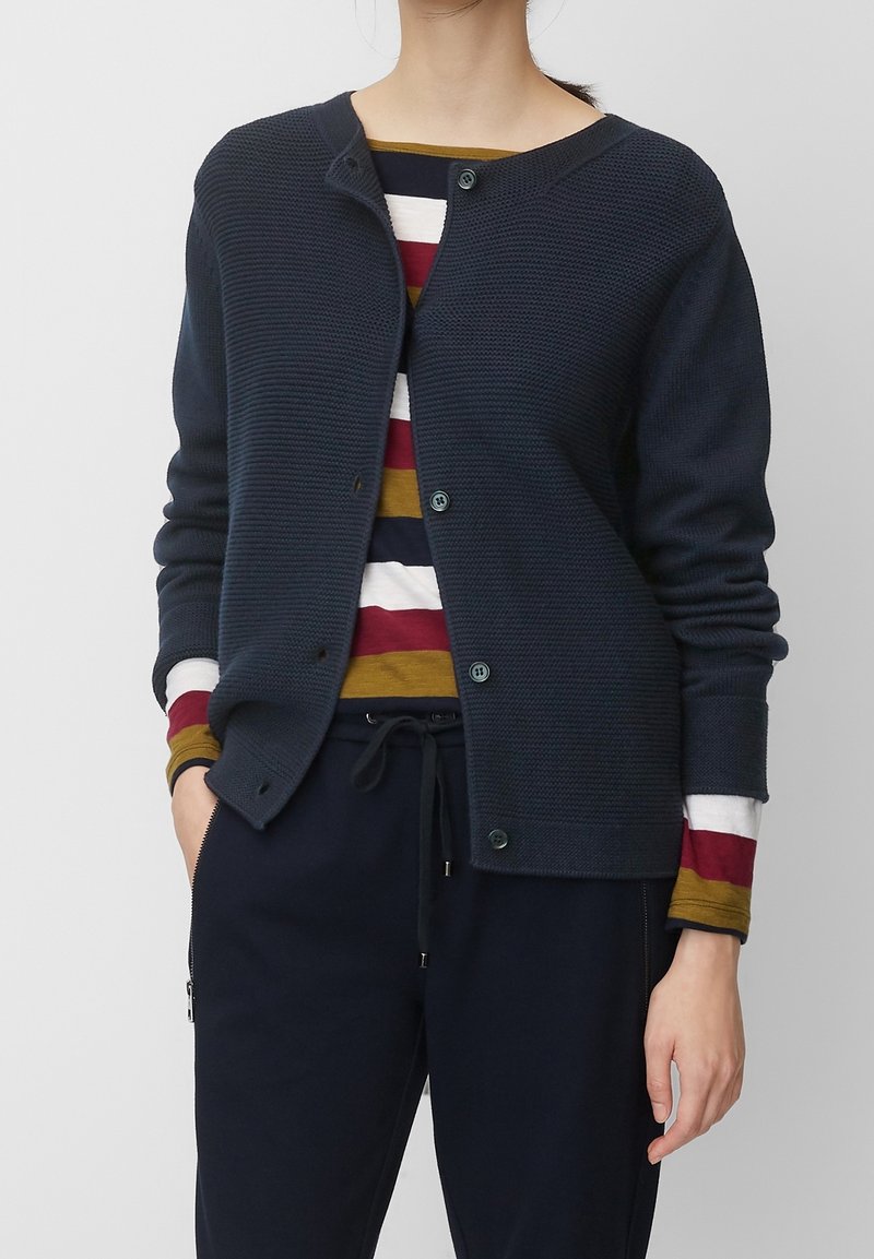 Femme portant un cardigan bleu marine boutonné par-dessus une chemise à manches longues rayée aux couleurs moutarde, bordeaux, blanc et bleu marine, et un pantalon bleu marine avec cordon de serrage.