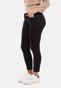 Persona che indossa jeans neri slim-fit, un maglione beige e sneakers bianche, in piedi con una mano in tasca su uno sfondo bianco.