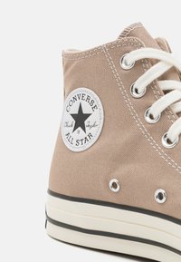 Baskets montantes en toile beige avec oeillets blancs, semelle en caoutchouc noire et patch logo circulaire avec un design d'étoile.