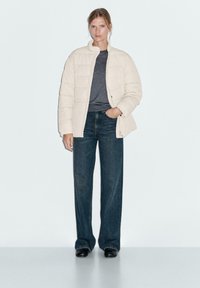 Femme portant une veste matelassée crème, un tee-shirt gris foncé, un jean large foncé et des chaussures noires, se tenant avec une main dans la poche.