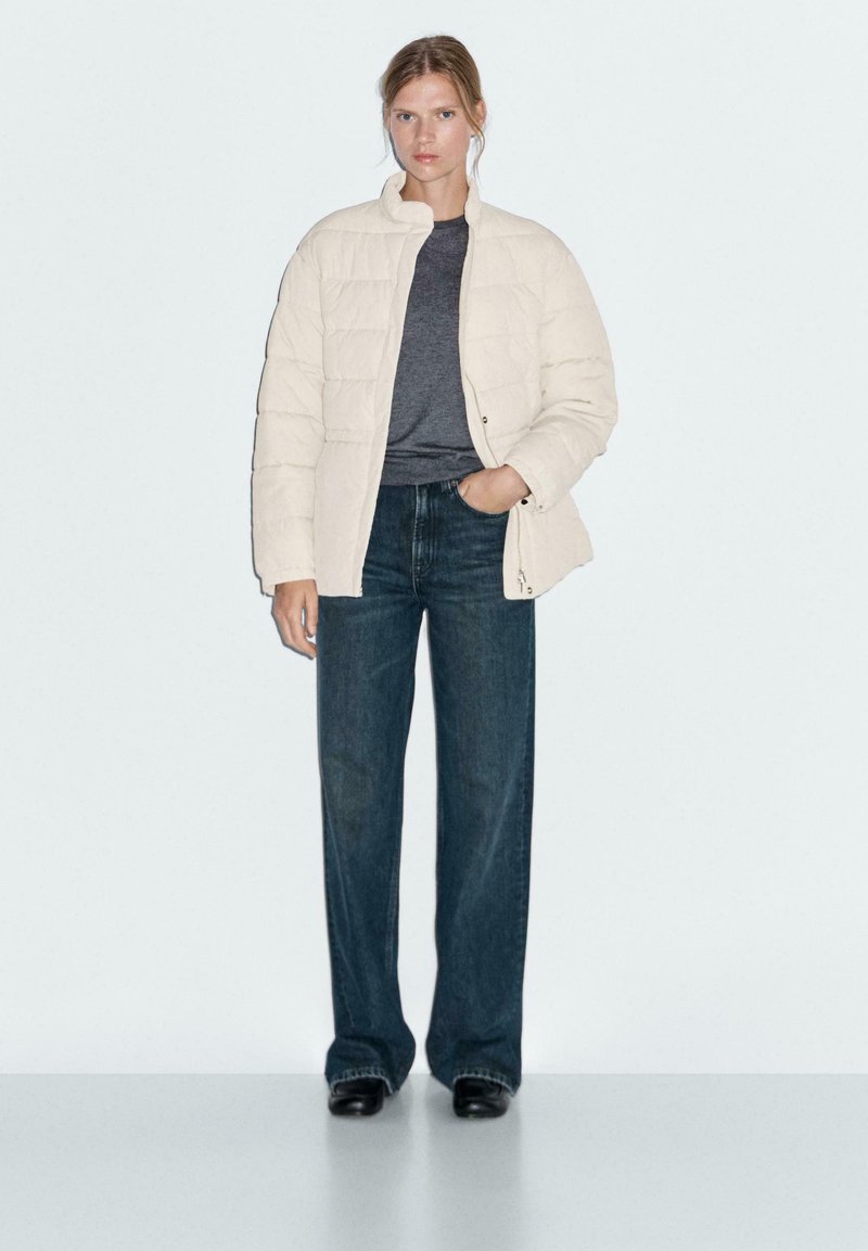 Femme portant une veste matelassée crème, un tee-shirt gris foncé, un jean large foncé et des chaussures noires, se tenant avec une main dans la poche.