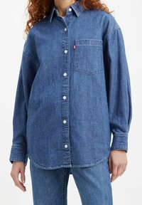 Chemise en denim bleu moyen avec col pointu, une poche poitrine unique, fermeture à boutons avec surpiqûres visibles et manches longues.
