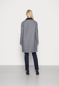 Donna in piedi di spalle che indossa un cappotto a scacchi bianco e nero, jeans blu scuro e scarpe con tacco nere su uno sfondo neutro.