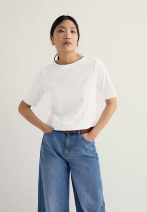 Fiatal nő fekete lófarokkal, arany karika fülbevalóval, fehér rövid ujjú ingben, barna övvel és széles kék farmerral, a kezét a zsebében tartva.