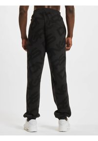 Svarte joggebukser med et stort logo-mønster, elastisk midje og en baklomme. Brukt sammen med hvite treningssko.