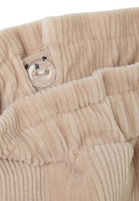 Beige corduroy stof met verticale ribbelstructuur en een zachte glans. Heeft een transparante knop en een geweven label met een logo.