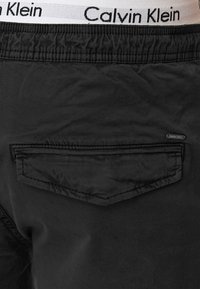 Shorts noirs avec une ceinture élastique et une poche arrière, arborant une bande de taille blanche visible de la marque Calvin Klein au-dessus des shorts.