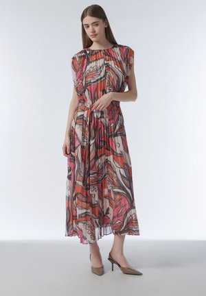 Young woman wearing sleeveless, pleated midi dress with abstract red, orange, and pink pattern, standing in neutral-toned heels against white backdrop.