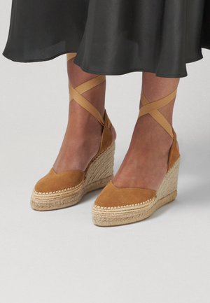 Bruine suède sleehakken met espadrille-zool en enkelbandjes, gedragen met een zwarte midirok op blote benen tegen een effen achtergrond.