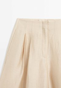 Jupe beige clair taille haute avec plis devant et fermeture de ceinture dissimulée sur fond blanc.