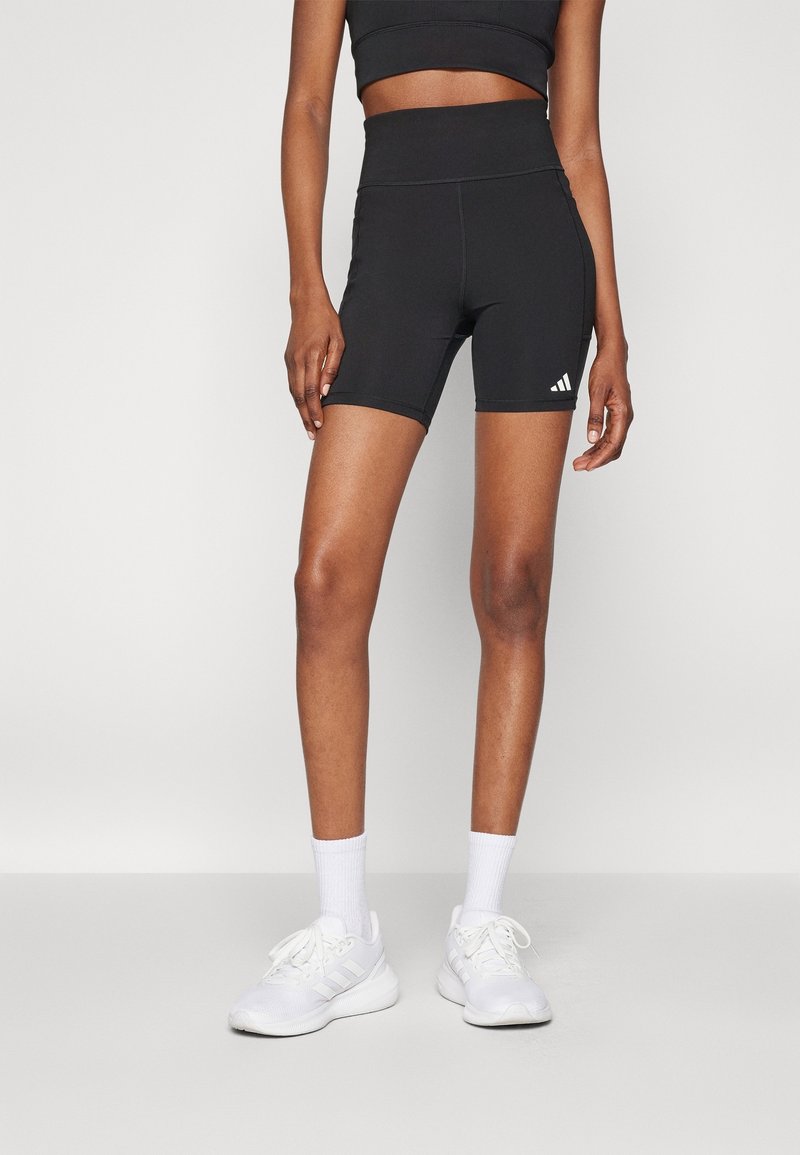 Shorts sportivi neri a vita alta, realizzati in materiale elastico, con un logo bianco sulla gamba sinistra. Abbinati a sneaker e calze bianche.