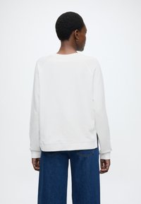 Fehér pulóver bordázott nyakkivágással és mandzsettákkal, raglán ujjakkal és oldalsó hasításokkal, széles szárú kék farmernadrággal párosítva.