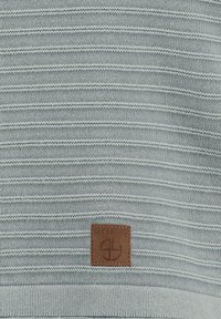 Maglione lavorato a maglia in blu-verdastro chiaro con strisce orizzontali. Presenta una toppa in pelle marrone nella parte inferiore con dettagli del logo. Texture morbida.