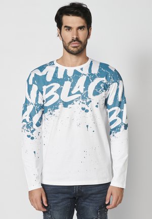 Langärmliges Shirt mit weißem Grundton, blauem Grafikdesign, auffälligem Text und Farbspritzern im oberen Bereich.