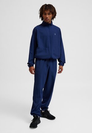 Navy Trainingsanzug aus glattem Stoff. Zip-Jacke mit Seitentaschen, passende Hose mit elastischem Bund und schwarze Sportschuhe.