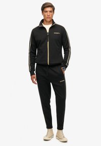 Man in een zwart Superdry trainingspak met beige strepen, beige hoge sneakers, een wit shirt en een polshorloge tegen een witte achtergrond.