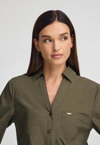 Olivgrön skjorta med knappar och krage, två bröstfickor samt en metallisk logotypaccents på vänster ficka. Mjuk och slät tyg.