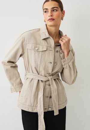 Woman wearing beige belted denim jacket with buttoned pockets, gold hoop earrings, and ring, looking to the side against a plain background.