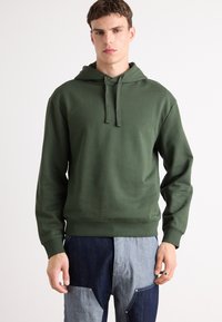 Felpa verde con cappuccio caratterizzata da una vestibilità comoda, tessuto in misto cotone e polsini e orlo a costine, indossata con pantaloni in denim con toppe.