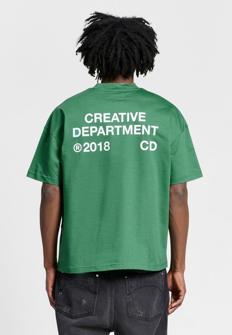 Camiseta verde de manga corta con texto blanco que dice "DEPARTAMENTO CREATIVO ® 2018 CD" en la parte de atrás. Material de algodón, corte relajado y sin patrones.