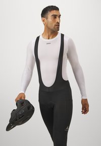 Schwarze Radsport-Bib-Shorts mit breiten Trägern über einem weißen langärmligen Mesh-Shirt. Die Person hält einen schwarzen Helm in der rechten Hand.