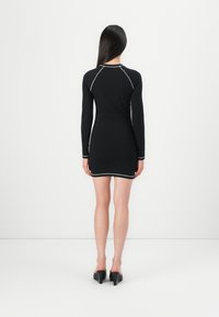 Robe noire à manches longues avec une silhouette ajustée, mettant en avant des coutures blanches contrastantes le long des coutures et de l'ourlet, associée à des chaussures à talons noires.