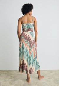 Maxi dress with thin straps, featuring a zigzag pattern in green, peach, and brown tones. Made of lightweight fabric, ankle-length with a fitted waist.