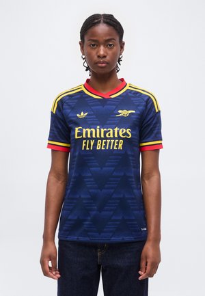 Ung person iført en blå og gul Arsenal fodboldtrøje med teksten "Emirates Fly Better", stående mod en ensfarvet hvid baggrund.