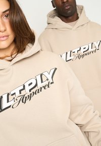 Felpe beige con cappuccio e lacci, caratterizzate da una grande grafica nera che recita "MULTPLY Apparel". Texture morbida, vestibilità rilassata e tasche a marsupio.
