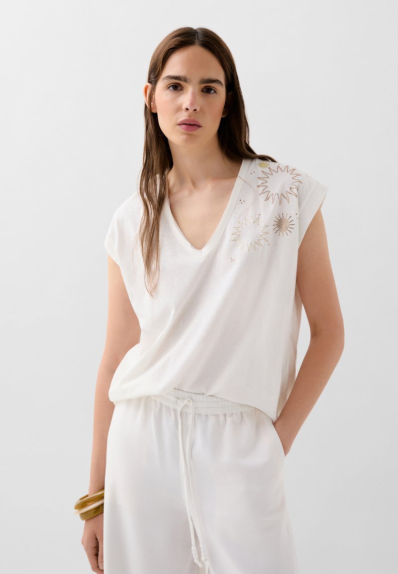White cotton v-neck top with short sleeves features embroidered sunburst patterns in gold and bronze on the left shoulder. Soft, relaxed fit.