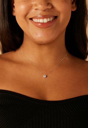 Collier en argent avec un pendentif rond étincelant sur une chaîne fine, porté sur une épaule nue devant un fond neutre.